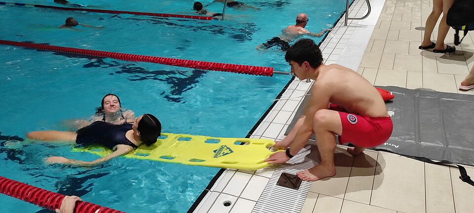 Eine Rettungsschwimmerin und ein Rettungsschwimmer üben im Hallenbad die Rettung einer Person aus dem Wasser. 